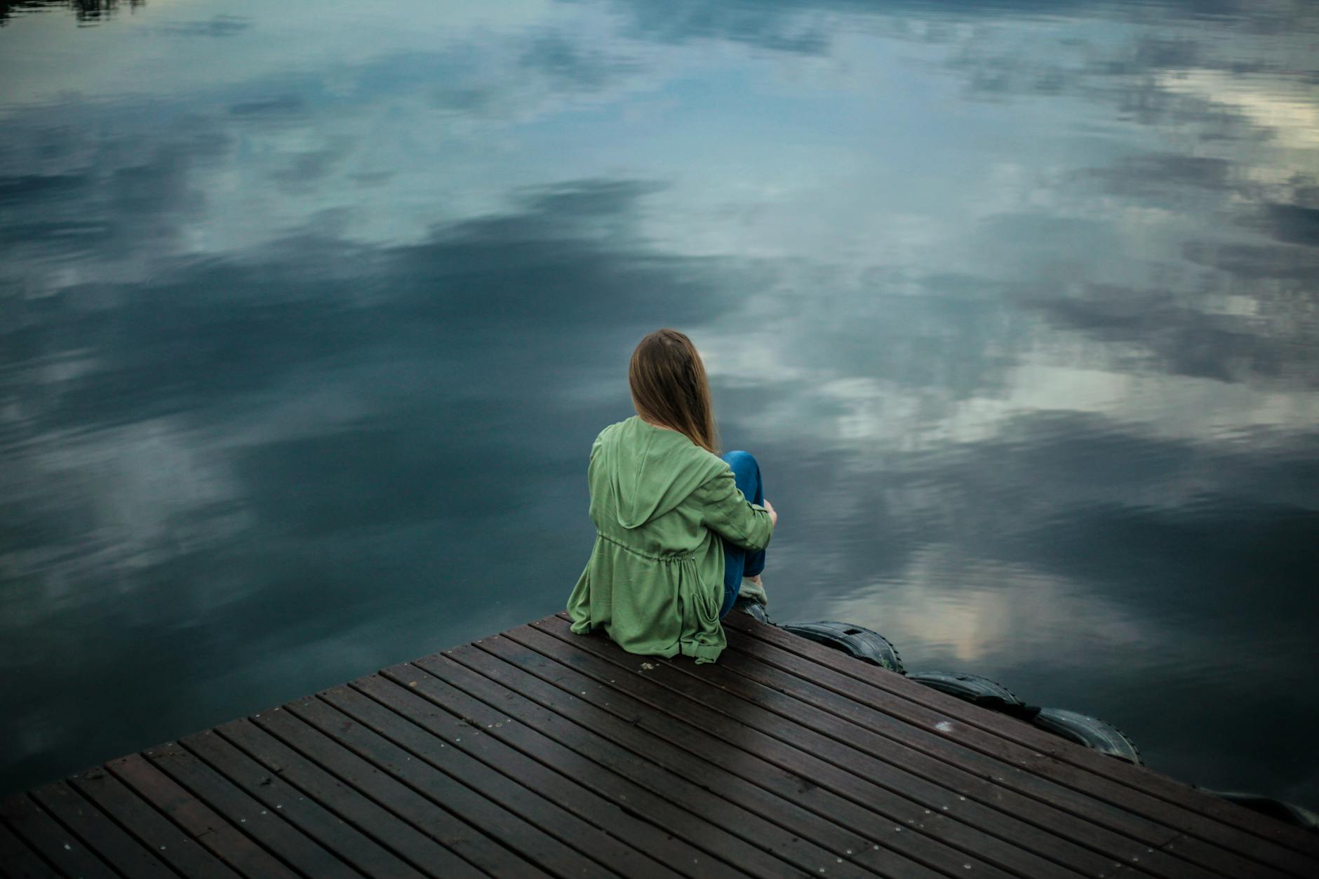 sad woman sitting alone at the edge of a pier on a lake on a gloomy day